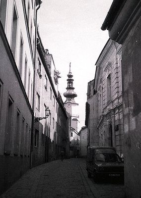 Narrow cobblestone alley flanked by historic European buildings, leading to a domed church tower. Mid-20th century vintage ca...
