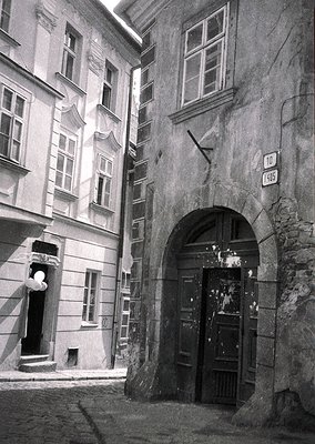 Historic European alleyway with weathered stone archway and cobblestone street. Multi-story buildings feature classical facad...