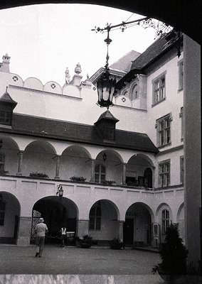 Neoclassical courtyard with arched colonnades, vintage streetlamp, and decorative rooftop elements. Two figures—one in light-...