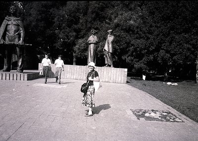 Black-and-white street scene featuring three prominent bronze statues of laborers in mid-gesture atop a raised platform, like...