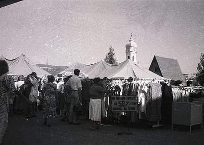 Outdoor market scene with clothing stalls under white tents, featuring a sign in Cyrillic advertising 50% discounts. A domed ...