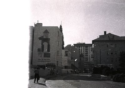 Mid-20th century urban street corner featuring a mix of Soviet-era architecture. Prominent building with Cyrillic signage rea...