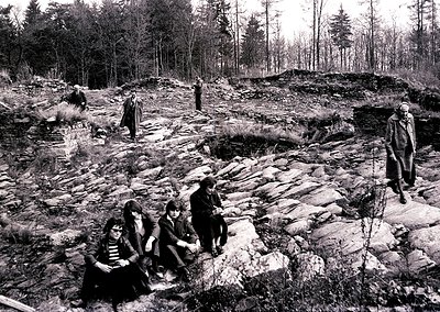 Black-and-white photo of a group exploring a rocky archaeological site in a forested area, likely Eastern Europe, 1960s-70s. ...