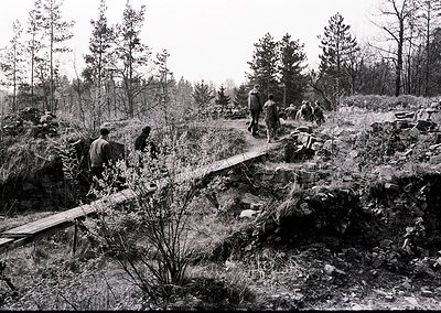 Group of men in military uniforms clearing a makeshift path through dense forest terrain, likely during WWII. Logs and branch...