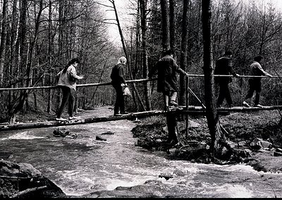 Four individuals cross a wooden suspension bridge over a shallow, rocky stream in a forested area, likely mid-20th century. B...