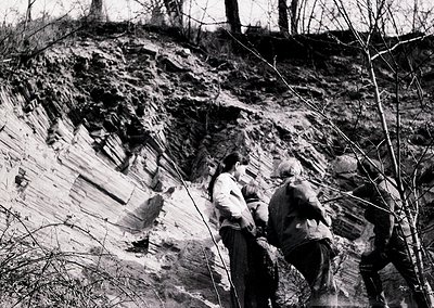 Two men examine exposed geological strata in a rocky cliffside, likely during mid-20th century. Distinct sedimentary layers a...