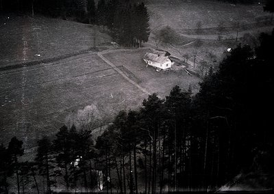 Aerial black-and-white photo of a rural homestead surrounded by dense forest. Single-story wooden house with a small yard, ad...