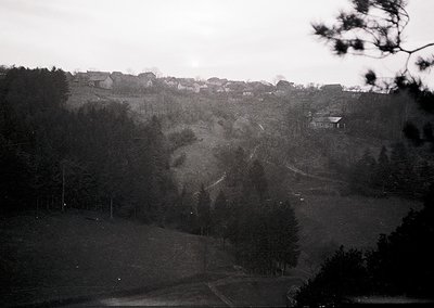 Vintage black-and-white landscape featuring a rural hillside settlement with scattered houses and dense forest. Rolling terra...