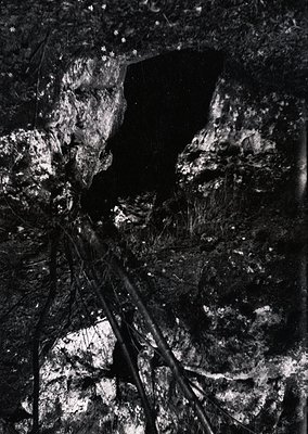 High-contrast black-and-white shot of a narrow, vertical cave entrance framed by jagged rock formations. Minimal light reveal...