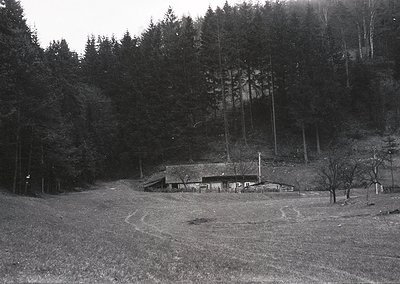 Black-and-white rural scene featuring a modest stone building with a pitched roof, surrounded by a fenced area. Dense forest ...
