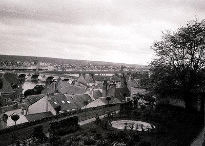 Mid-20th century European town from elevated vantage point, showcasing dense rooftops, a stone bridge, and a circular fountai...