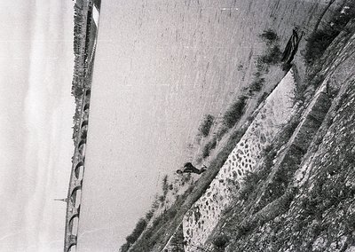 Aerial black-and-white photo of a steep, terraced hillside with stone retaining walls. A lone figure in dark clothing appears...
