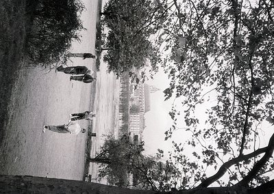 Vintage black-and-white shot of a seaside promenade with three figures in mid-stride, framed by leafy trees. The curved concr...