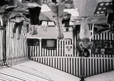 Vintage black-and-white photo of a playful indoor scene. Three individuals upside-down on striped bedsheets, suspended by a m...