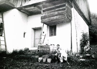 Traditional wooden house exterior with elevated balcony, likely rural Eastern Europe. Woman seated on steps, dressed in long-...