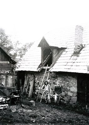 Rural stone cottage with wooden roof and exposed beams, likely Eastern European. Two men pose near a ladder and wooden cart, ...