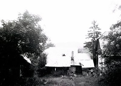 Early 20th-century farmstead with weathered wooden structures and overgrown vegetation. Sloped roof barn and smaller outbuild...