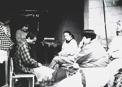 Group of five individuals engaged in textile work in a dimly lit, rustic indoor setting, likely a workshop or communal space....