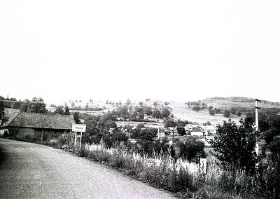 Rural village roadside scene with modest brick homes, overgrown vegetation, and a hand-painted "Varna" sign. Mid-20th century...