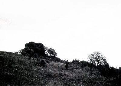 Black-and-white landscape featuring lone figure ascending a grassy hill toward a prominent rock formation. Mid-20th century o...