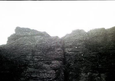 Black-and-white shot of rugged, layered rock formations under misty skies. Dramatic vertical cliffs and horizontal strata sug...