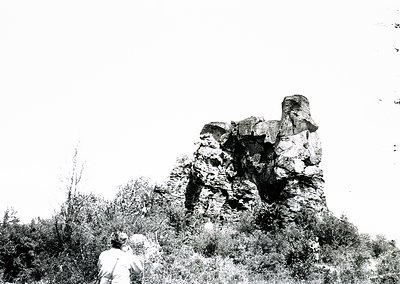 Black-and-white photograph of a lone figure examining a large, weathered rock formation resembling a seated figure or throne....