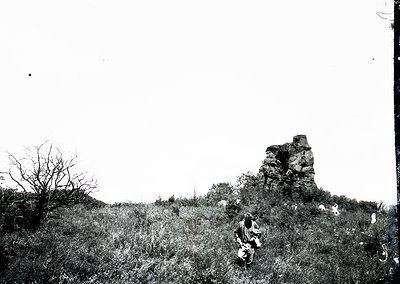 Vintage black-and-white photo of a lone figure seated atop a grassy hillside, with a prominent rock formation in the backgrou...
