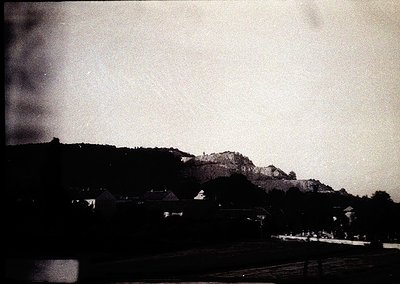 Vintage black-and-white landscape featuring a low-contrast rural scene with dense forest and rocky hillside. Small, clustered...