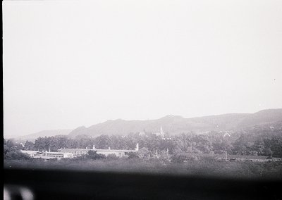 Vintage black-and-white landscape featuring a low-angle view of a large, symmetrical institutional building surrounded by den...
