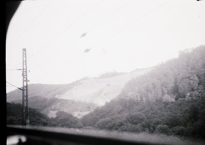 Black-and-white train window view of misty, snow-capped alpine ridge. Power lines and forest below suggest mid-20th century E...
