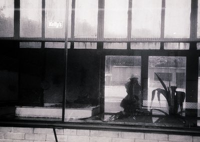 Black-and-white street scene captured through a storefront window, featuring a seated figure in a wide-brimmed hat and dark c...
