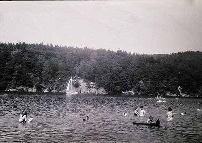 Vintage black-and-white seaside scene with 1950s-60s swimwear, including men in trunks and women in one-piece suits. Group of...