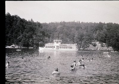 Vintage black-and-white photo of a mid-20th century lakeside scene. A group of people swim in calm waters near a docked ferry...