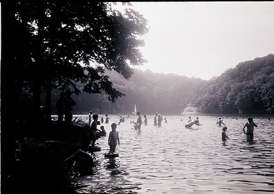 Black-and-white scene of a lakeside gathering, likely mid-20th century. Dense tree canopy frames a shallow lake where childre...