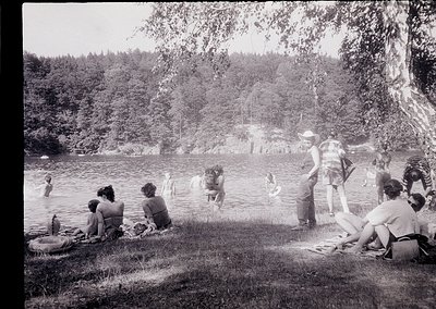 Mid-20th century lakeside gathering with 1950s–60s swimwear and vintage floatation devices. Group of men and women lounging o...