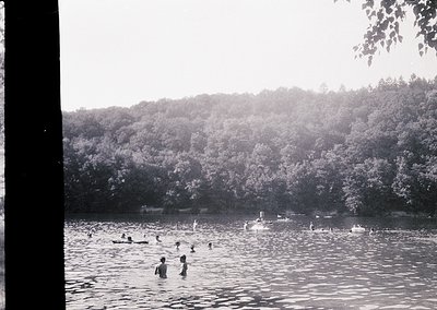 Vintage black-and-white photo of a crowded natural swimming area with dense forest backdrop. Mid-20th century attire (swimsui...
