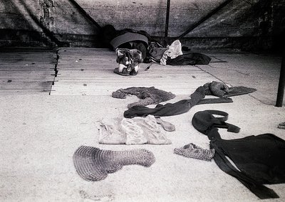 Black-and-white photo of discarded clothing and textiles on a concrete floor, likely a basement or storage area. Items includ...