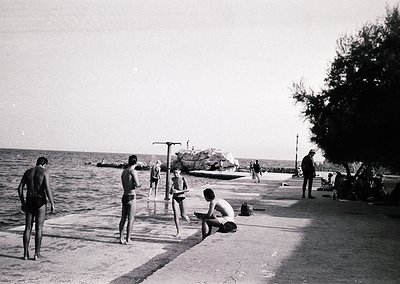 Black-and-white seaside scene with young men in swim trunks (1950s–60s style) by a concrete pier. One man sits reading, other...