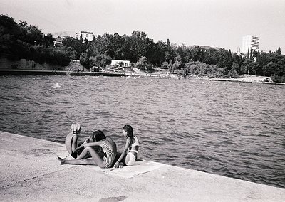 Three women in 1960s-style swimsuits sit on a concrete ledge by a lake, backs to camera. Mid-century urban park in background...