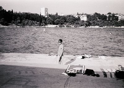 Mid-20th century seaside scene: a lone child in shorts stands on a sandy shore, framed by scattered towels and a rolled blank...