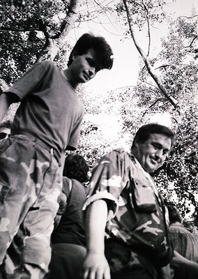 Black-and-white candid of two men laughing while hiking, framed by dense forest foliage. Mid-20th century outdoor attire sugg...