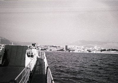 Black-and-white coastal scene featuring a ferry’s bow with a lone figure mid-dive into the sea. Urban coastline with mid-rise...
