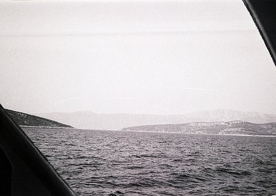 Black-and-white seascape shot from a boat’s perspective, showing choppy waters and distant landmass with faint outlines of tr...