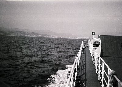 Black-and-white shot of a ferry’s bow cutting through calm waters, with distant coastal hills and misty horizon. Mid-20th cen...