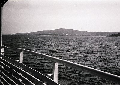 Black-and-white seascape shot from a ship’s railing, showing calm waters and distant hilly coastline. Mid-20th century mariti...