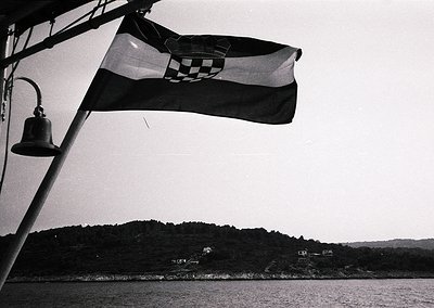 Croatian flag waves atop a ship’s mast beside a handbell, set against a calm coastal landscape. Black-and-white monochrome su...