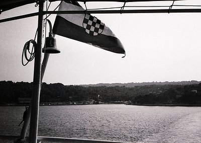 Croatian flag with checkerboard pattern waves over calm waters. Bell and rope detail on ship’s rail. Dense forested coastline...