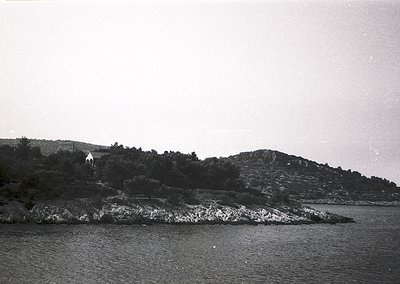 Black-and-white coastal scene featuring jagged rocky shoreline and calm waters. A lone figure stands atop a hillside covered ...