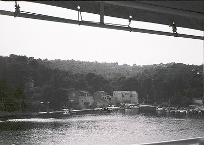 Black-and-white coastal scene featuring a bridge with hanging light fixtures framing the view. Below, a serene harbor with bo...