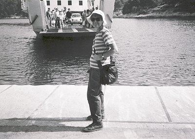 Mid-20th century ferry scene: Woman in striped blouse, wide-brim hat, and bag stands on concrete pier beside calm water. Clas...